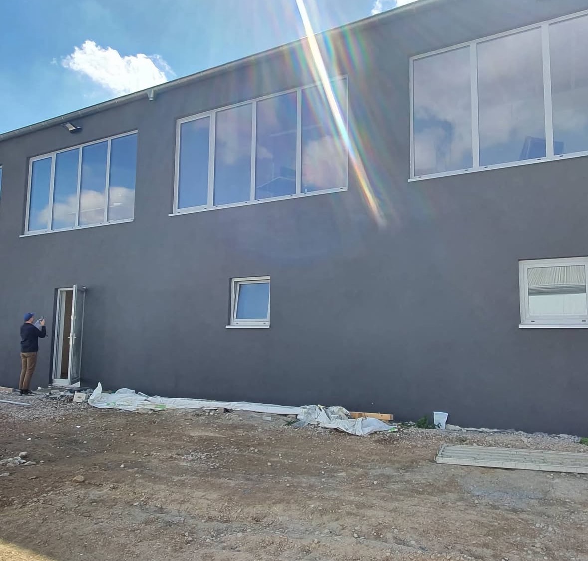 Szara elewacja budynku z białymi oknami. Osoba robi zdjęcie przy otwartych drzwiach. Widoczne materiały budowlane i niezagospodarowany teren przed budynkiem.