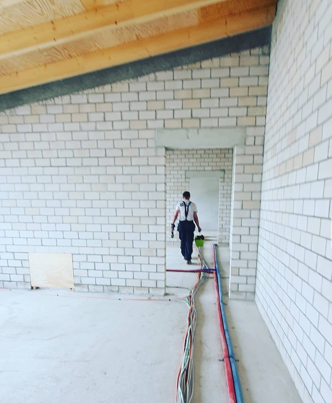 Wnętrze budynku w trakcie budowy: murowane ściany z białej cegły, drewniany strop i widoczne instalacje (kable, rury) na podłodze. W tle sylwetka osoby.