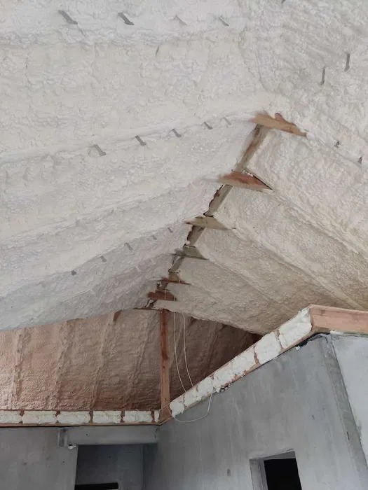 Poddasze z widoczną warstwą białej pianki izolacyjnej, drewnianą konstrukcją i elementami mocującymi. Częściowo ocieplona ściana szczytowa. Surowy stan.