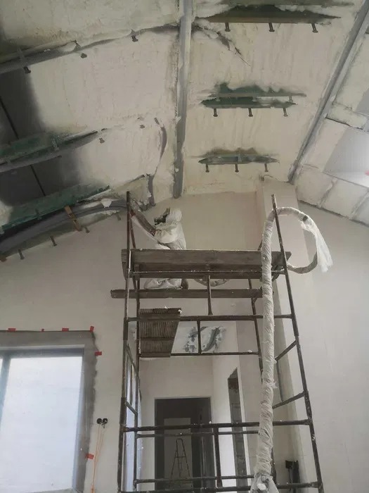 Prace izolacyjne pianką PUR na poddaszu: pracownik w kombinezonie na rusztowaniu aplikuje pianę na skośny sufit z widocznymi elementami konstrukcyjnymi.