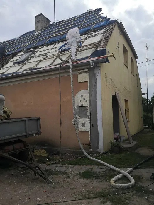 Prace na dachu: aplikacja izolacji pianką na odsłoniętej konstrukcji. Pracownik w kombinezonie ochronnym, przewód doprowadzający pianę, fragment elewacji budynku.