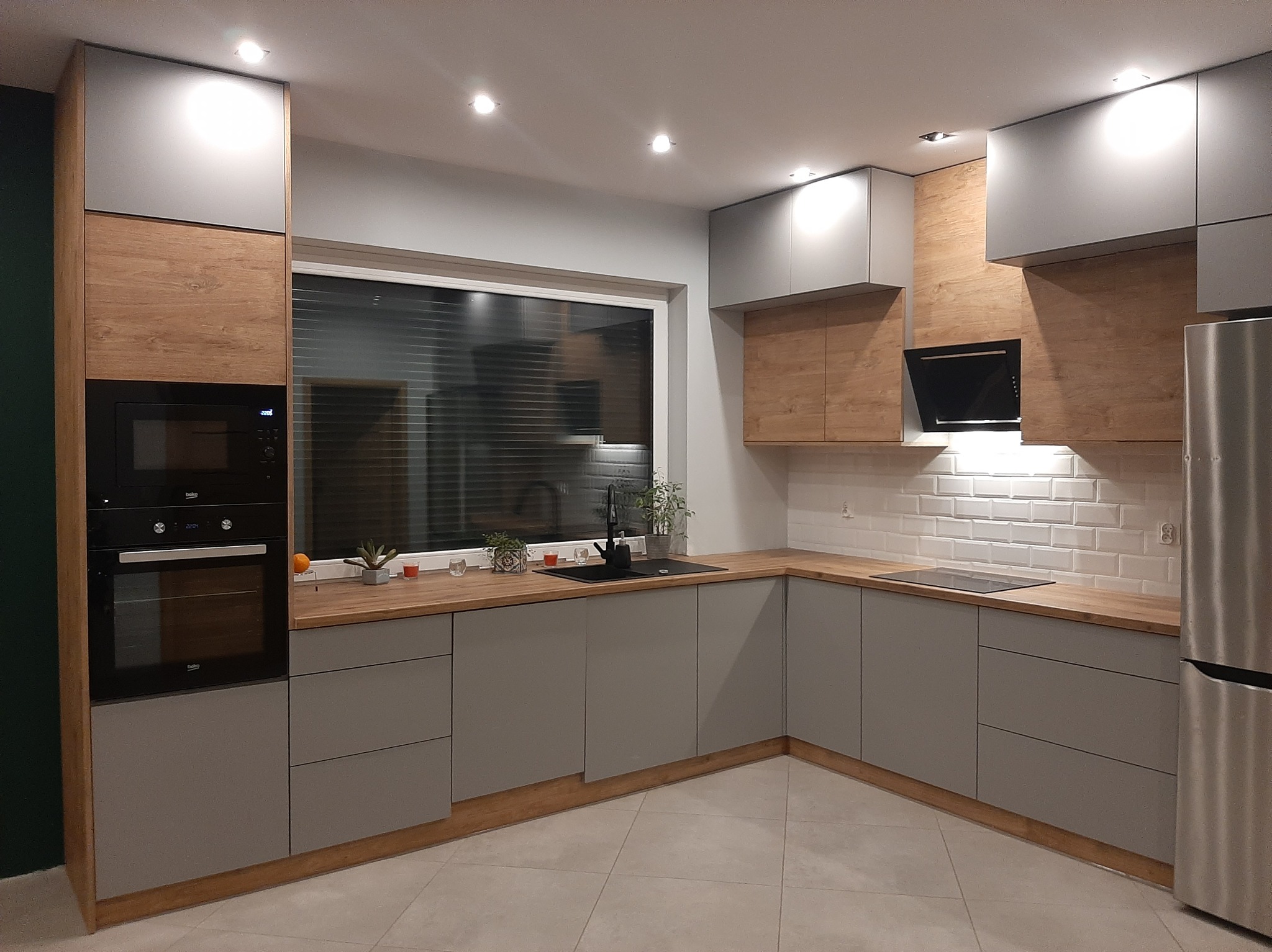 Nowoczesna kuchnia z szarymi szafkami i drewnianymi akcentami, oświetleniem punktowym i białymi płytkami metra na ścianie. Minimalistyczny design z czarną armaturą.