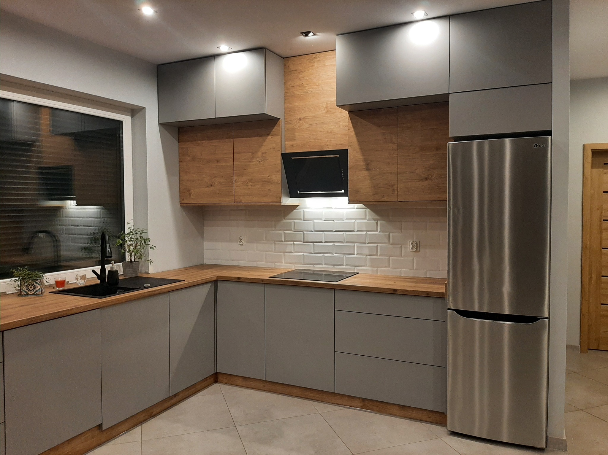 Nowoczesna kuchnia: szare szafki, drewniane akcenty, białe kafelki. Uwagę przyciąga srebrna lodówka LG i czarny okap. Oświetlenie punktowe podkreśla detale.