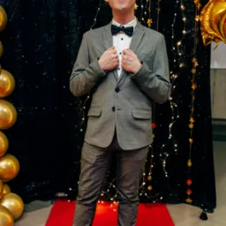 Mężczyzna w szarym garniturze i zielonych okularach pozuje na czerwonym dywanie z dekoracjami świetlnymi i złotymi balonami. Uśmiechnięty, elegancki strój.