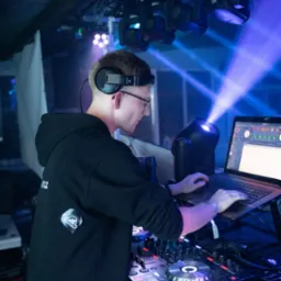 DJ z Oławy w trakcie występu, skupiony na miksowaniu muzyki. Widoczne oświetlenie sceniczne i sprzęt DJ-ski Pioneer. Ujęcie z boku, podkreślające profesjonalizm.