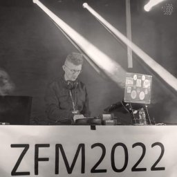 Kamil Żmijowski - DJ Kamz - Czarno-białe zdjęcie DJ-a w trakcie występu na scenie, oświetlonej reflektorami. Na stole widoczny napis 'ZFM 2022'. DJ ma słuchawki na szyi i skupia się na miksowaniu muzyki.