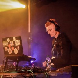 Kamil Żmijowski - DJ Kamz - DJ z rudymi włosami w okularach, w słuchawkach, miksuje muzykę na konsolecie. Laptop z naklejkami, oświetlenie sceniczne w tle. Butelka wody obok sprzętu.