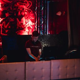 Kamil Żmijowski - DJ Kamz - DJ w czerwonym oświetleniu, skupiony na miksowaniu muzyki na konsoli. W tle dekoracje i publiczność, tworzące klubową atmosferę. Widoczny laptop i słuchawki.