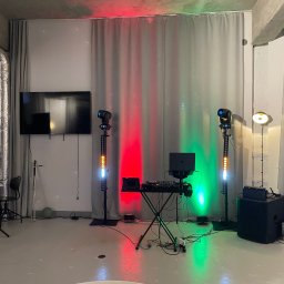 Kamil Żmijowski - DJ Kamz - Sprzęt DJ-ski z laptopem, mikserem i oświetleniem scenicznym w kolorach czerwonym i zielonym, ustawiony na tle szarych zasłon i betonowego sufitu.