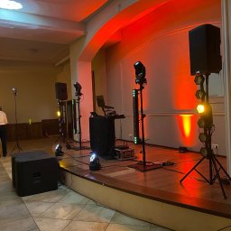 Kamil Żmijowski - DJ Kamz - Scena z nagłośnieniem i oświetleniem gotowa na występ. Widoczne kolumny głośnikowe, reflektory i konsoleta. W tle osoba w jasnej koszuli. Scena oświetlona na czerwono.