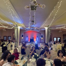 Kamil Żmijowski - DJ Kamz - Sala balowa z oświetleniem dekoracyjnym i gośćmi przy stołach. Scena z nagłośnieniem w tle. Dekoracje świetlne tworzą ciepłą atmosferę.