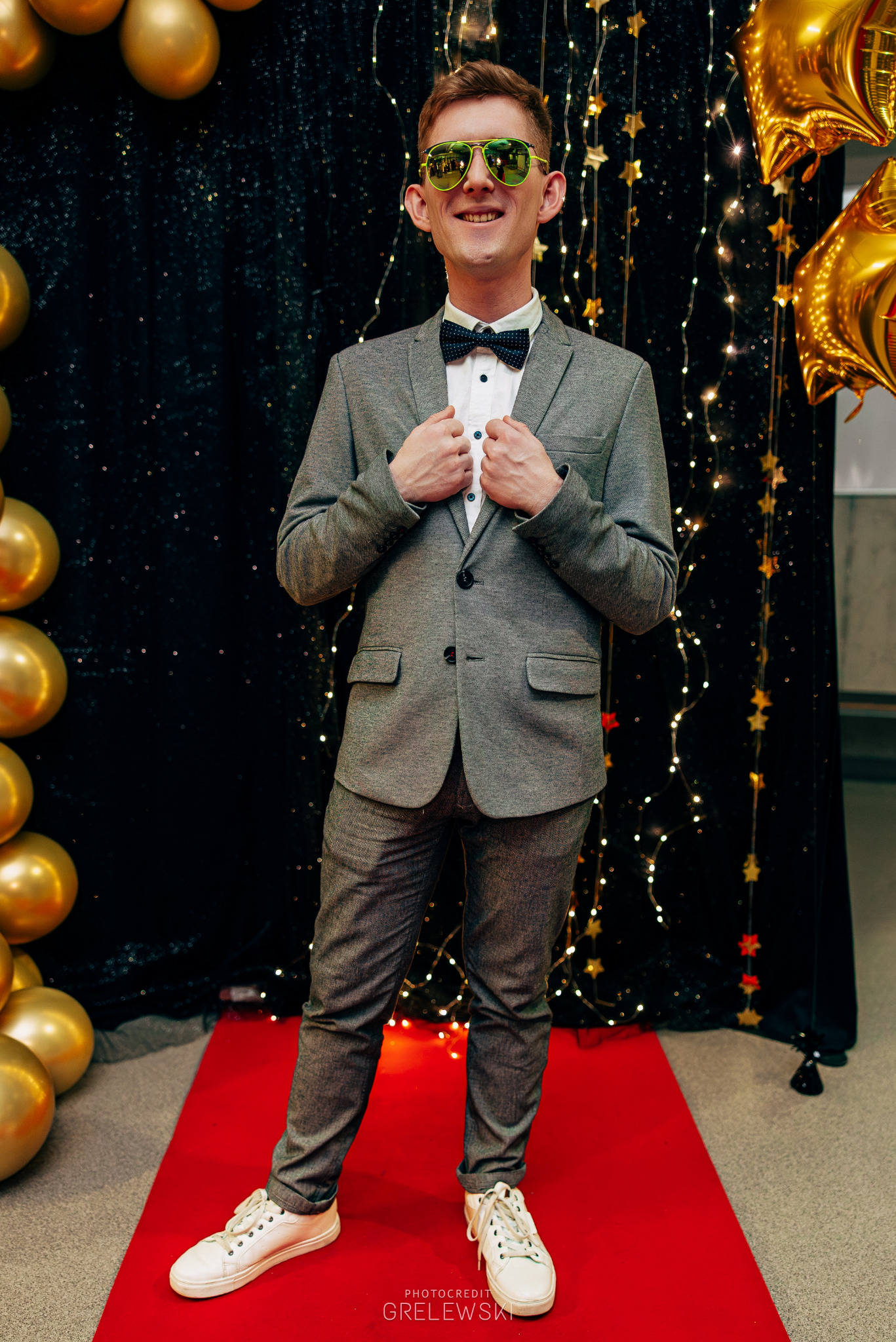 Mężczyzna w szarym garniturze i zielonych okularach pozuje na czerwonym dywanie z dekoracjami świetlnymi i złotymi balonami. Uśmiechnięty, elegancki strój.