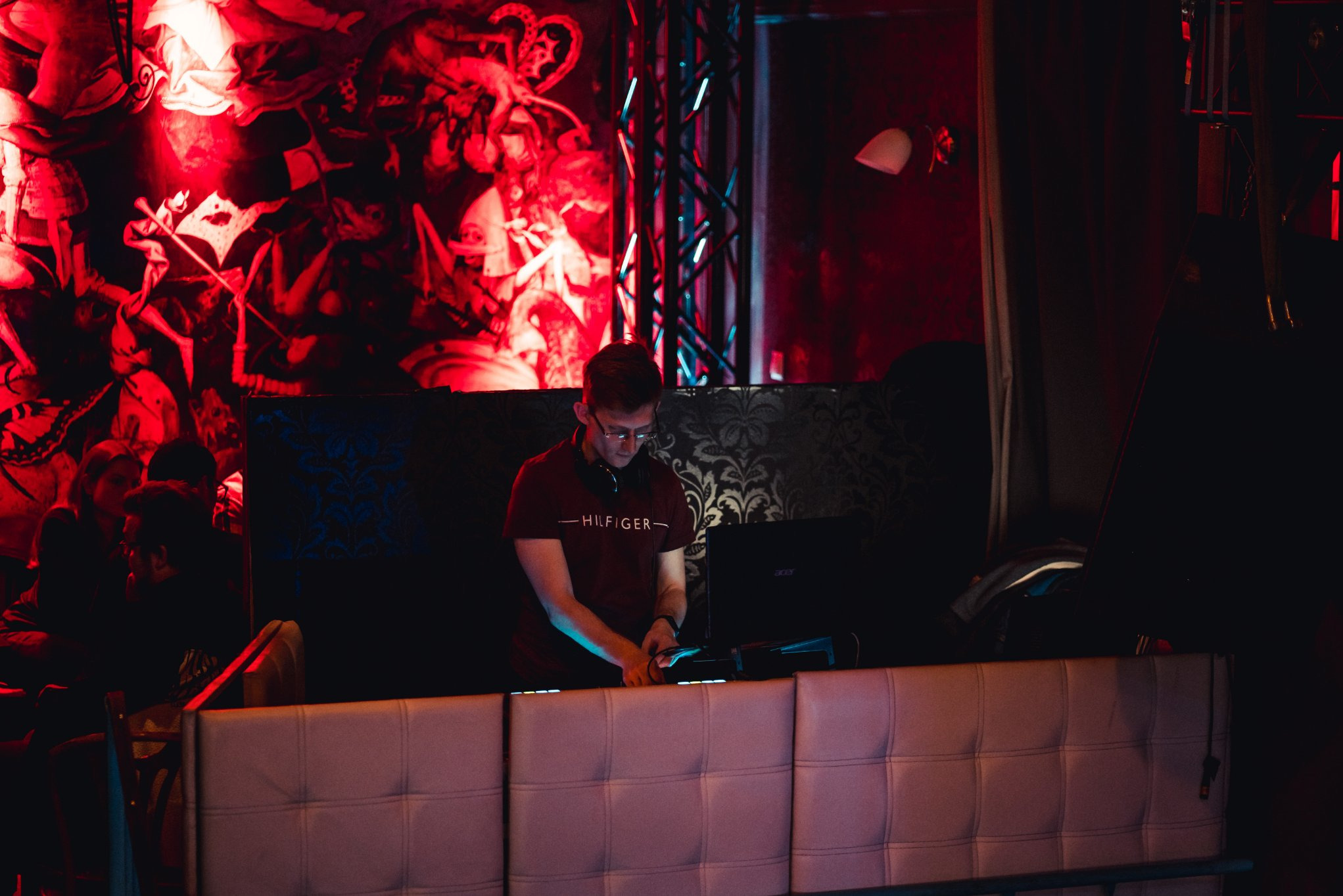 DJ w czerwonym oświetleniu, miksujący muzykę na konsolecie w klubie. Widoczny laptop i słuchawki. Dekoracje na ścianie i publiczność w tle.