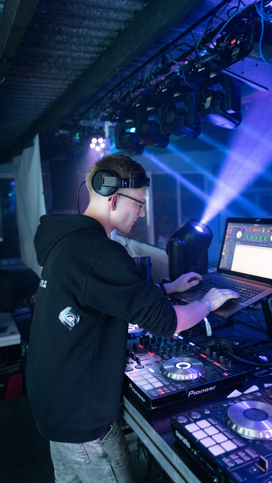 DJ z Oławy w trakcie występu, skupiony na miksowaniu muzyki. Widoczne oświetlenie sceniczne i sprzęt DJ-ski Pioneer. Ujęcie z boku, podkreślające profesjonalizm.