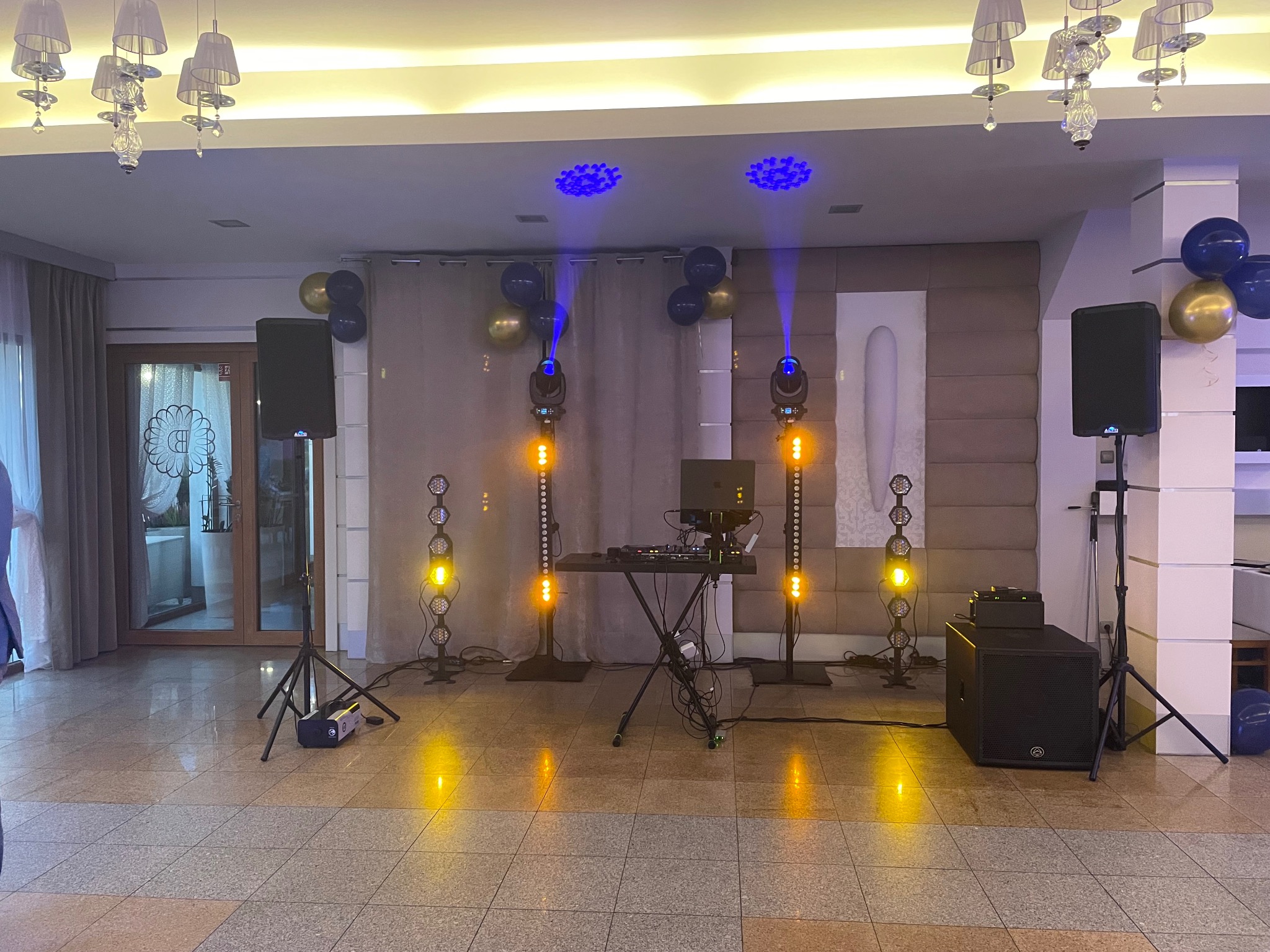 Sala bankietowa z nagłośnieniem i oświetleniem: kolumny głośnikowe, reflektory z niebieskim i pomarańczowym światłem, konsoleta DJ-ska. Dekoracja balonowa.