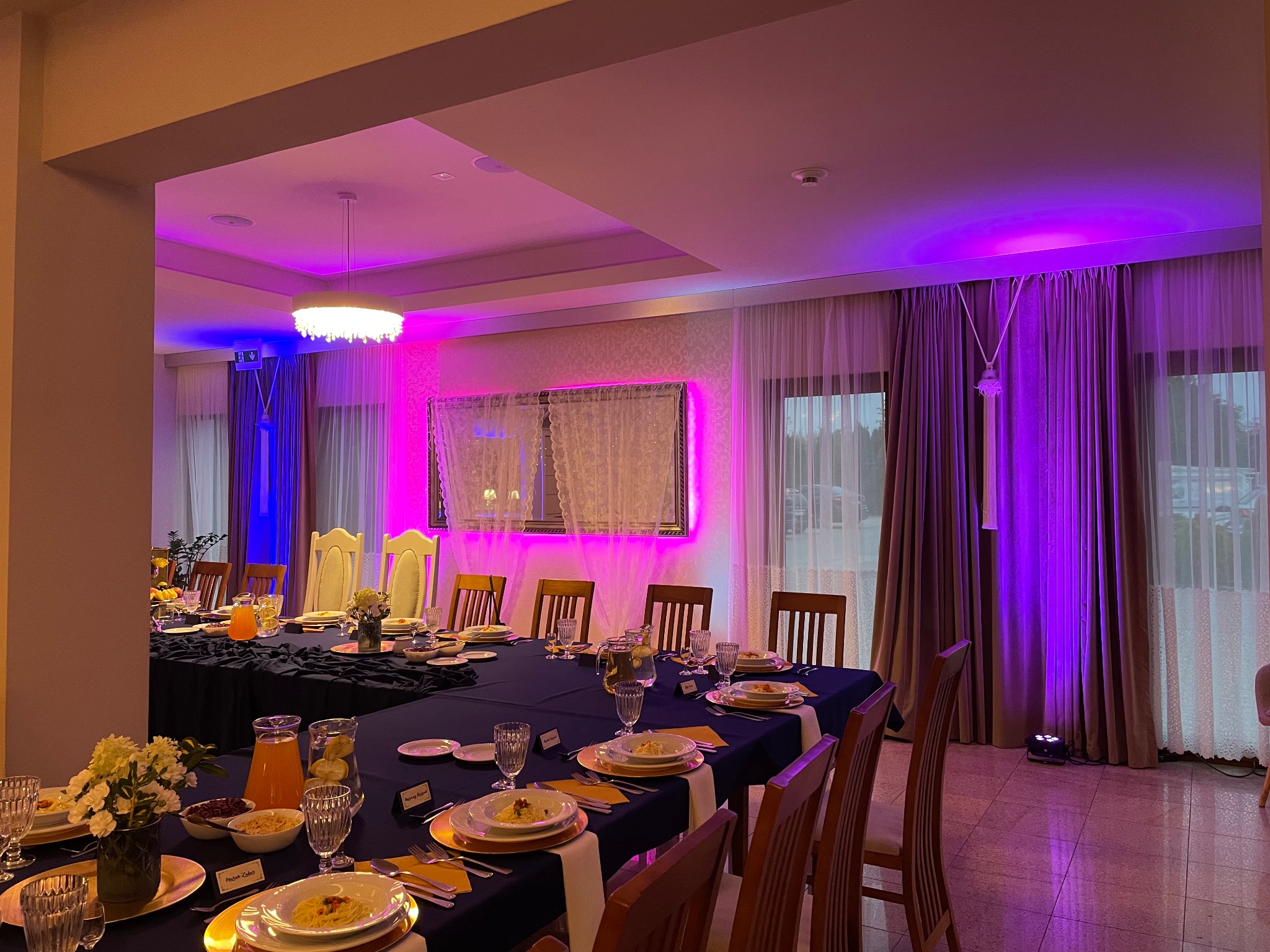 Uroczysty stół bankietowy z ciemnym obrusem, zastawą, kwiatami i dekoracyjnym oświetleniem w odcieniach fioletu i błękitu, przygotowany na przyjęcie.