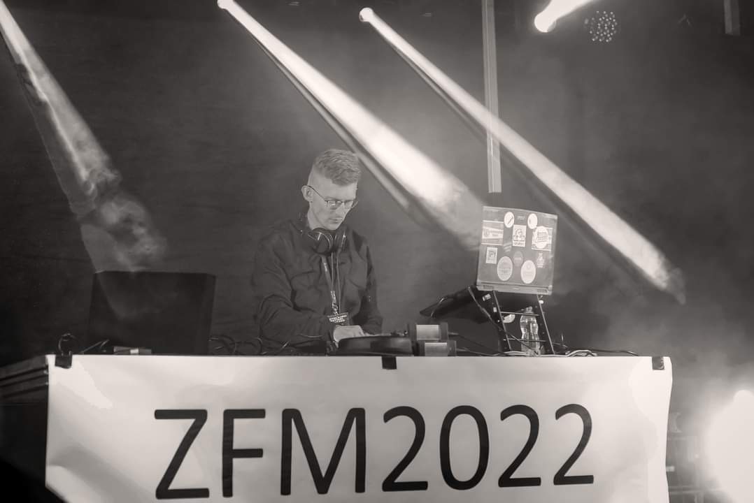 Czarno-białe zdjęcie DJ-a w trakcie występu na scenie, oświetlonej reflektorami. Na stole widoczny napis 'ZFM 2022'. DJ ma słuchawki na szyi i skupia się na miksowaniu muzyki.