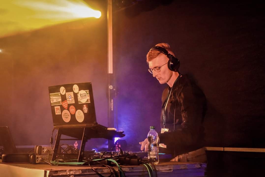 DJ z rudymi włosami w okularach, w słuchawkach, miksuje muzykę na konsolecie. Laptop z naklejkami, oświetlenie sceniczne w tle. Butelka wody obok sprzętu.