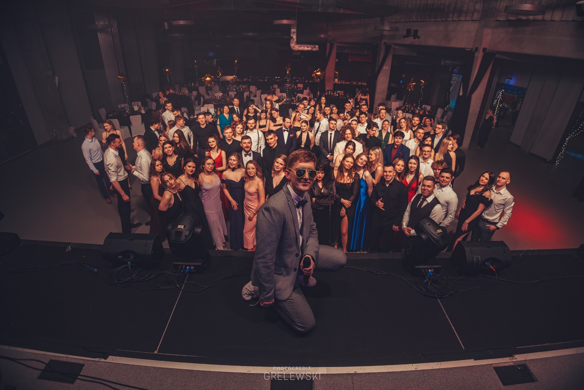 Szeroki kadr na grupę elegancko ubranych osób na scenie, z mężczyzną w szarym garniturze i okularach na pierwszym planie, oświetlenie sceniczne, uroczystość.