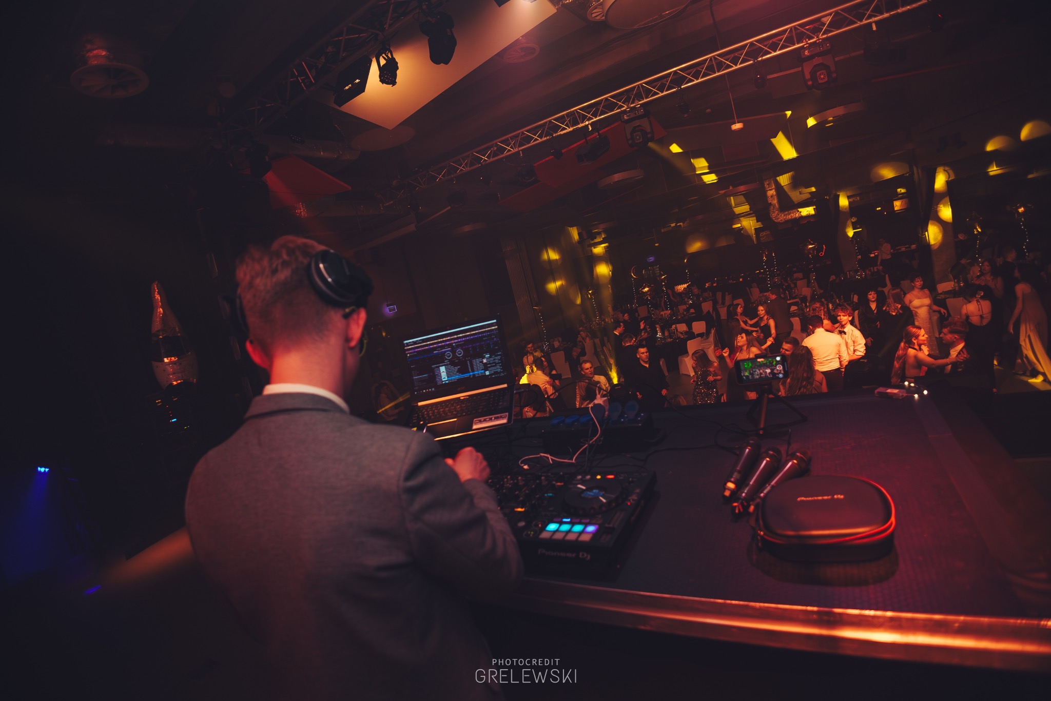 DJ w szarej marynarce miksuje muzykę na imprezie. W tle oświetlenie sceniczne i bawiący się ludzie. Widok zza pleców DJ-a, sprzęt Pioneer DJ.