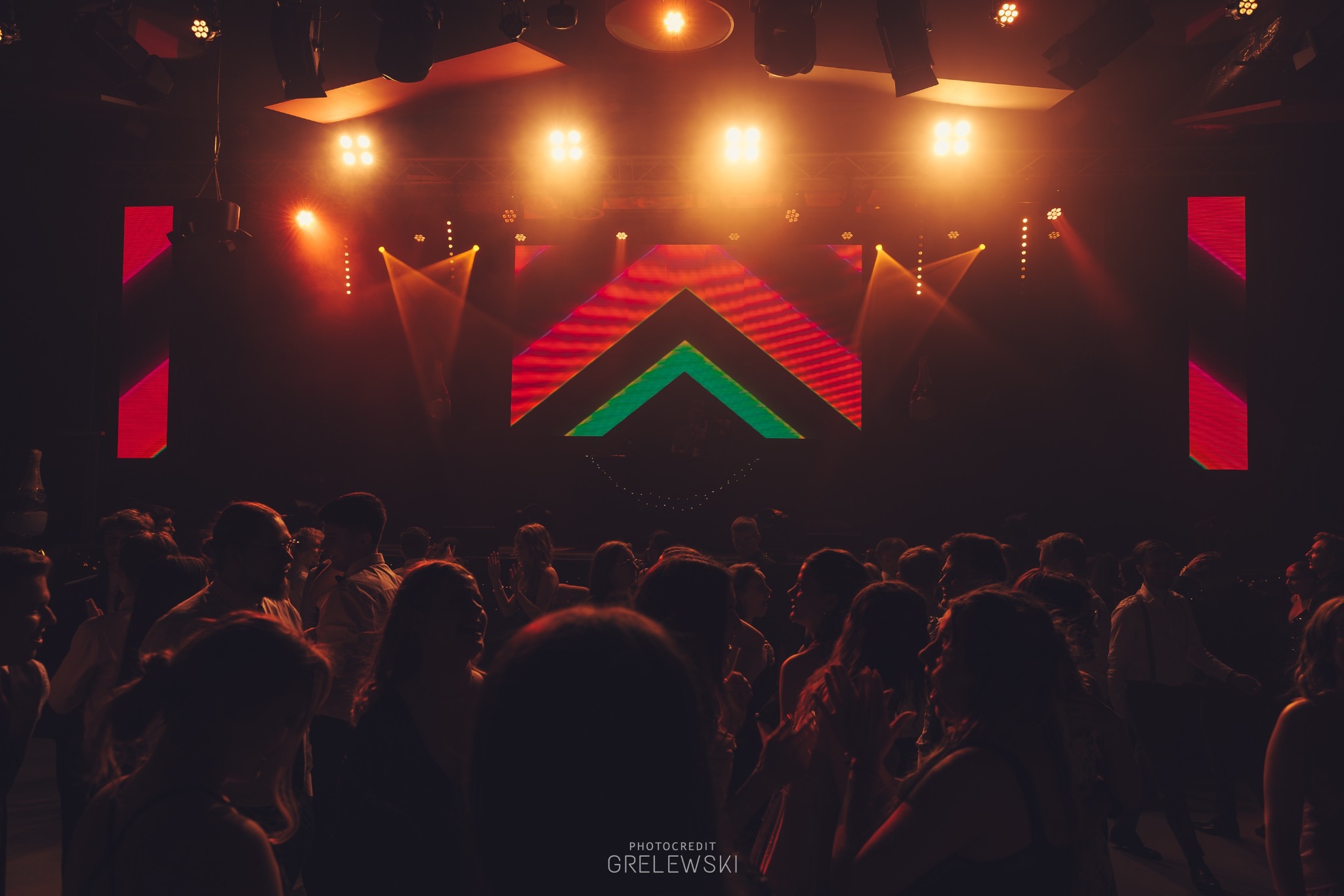 Tłum ludzi w ciemnym klubie nocnym, oświetlonym przez reflektory i ekran LED z geometrycznym wzorem. Widoczny parkiet i elementy nagłośnienia.