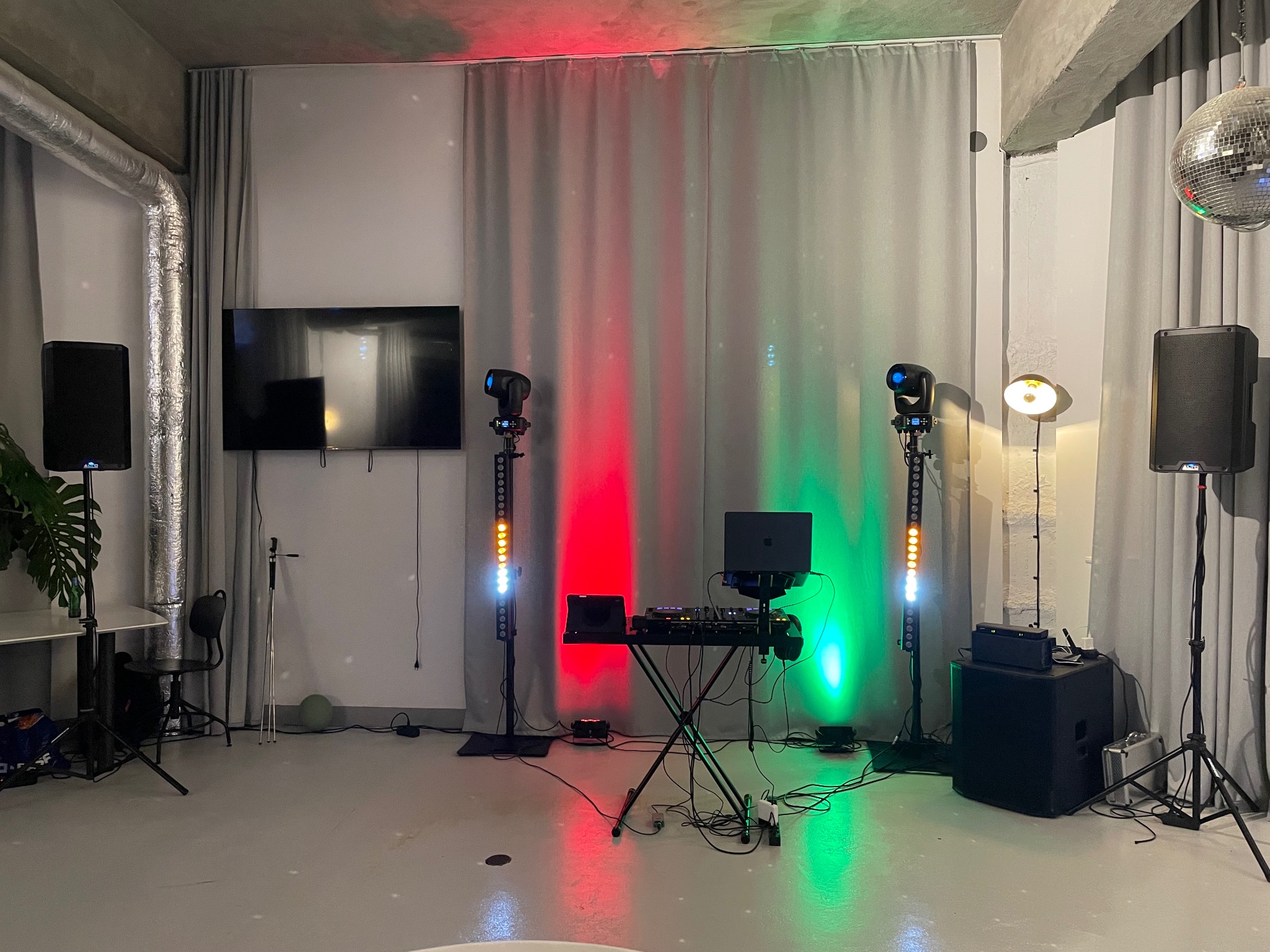 Sprzęt DJ-ski z laptopem, mikserem i oświetleniem scenicznym w kolorach czerwonym i zielonym, ustawiony na tle szarych zasłon i betonowego sufitu.