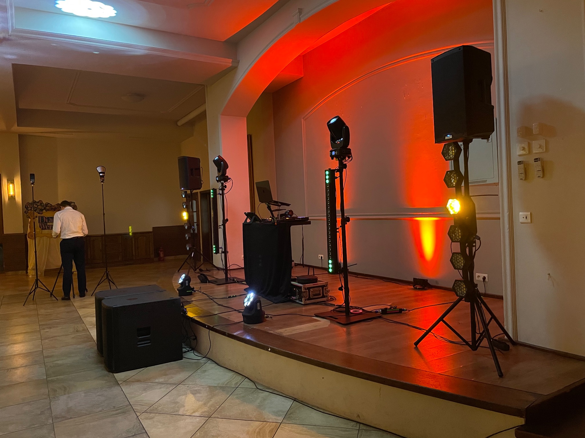 Scena z nagłośnieniem i oświetleniem gotowa na występ. Widoczne kolumny głośnikowe, reflektory i konsoleta. W tle osoba w jasnej koszuli. Scena oświetlona na czerwono.