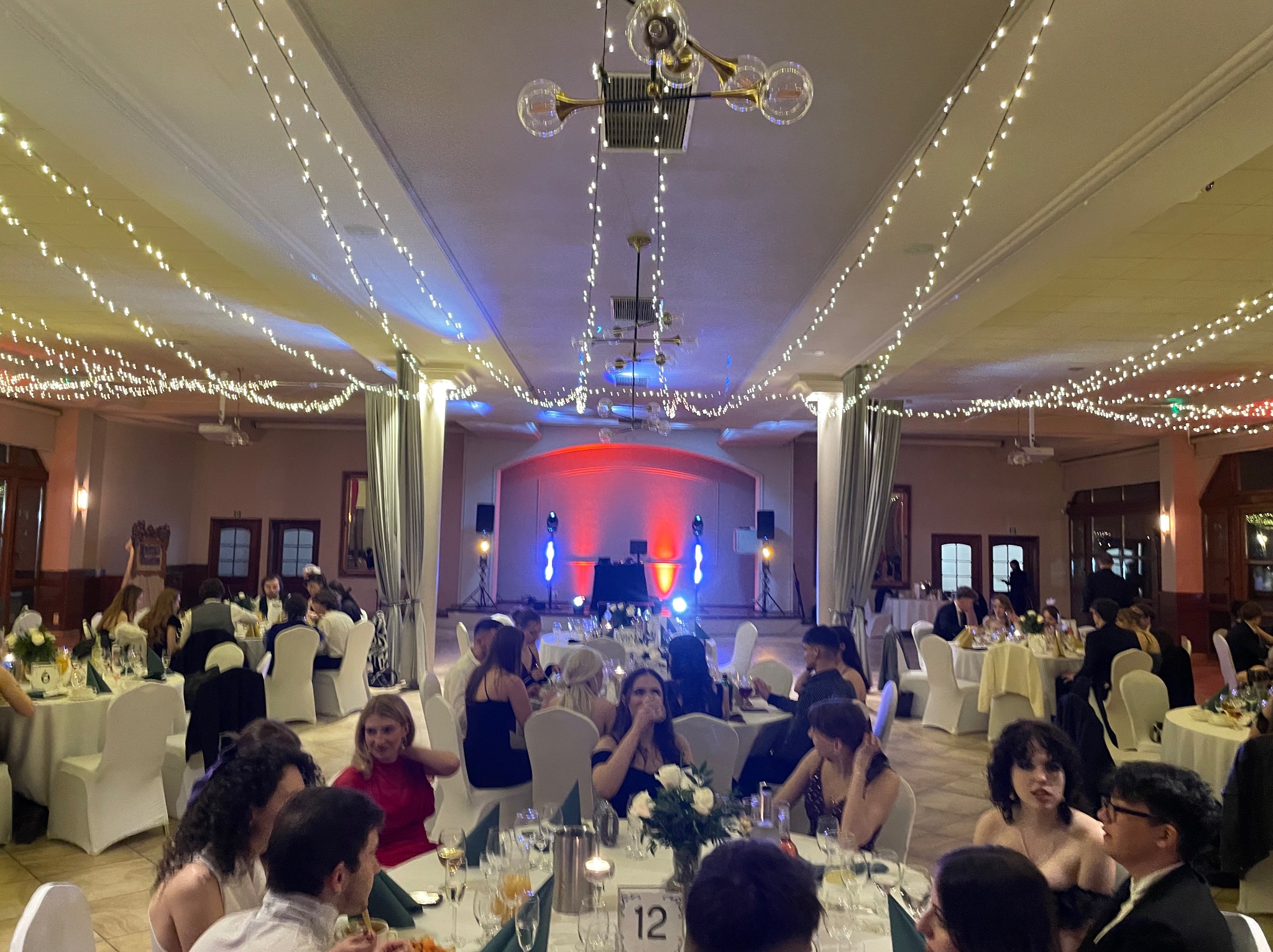 Sala balowa z oświetleniem dekoracyjnym i gośćmi przy stołach. Scena z nagłośnieniem w tle. Dekoracje świetlne tworzą ciepłą atmosferę.