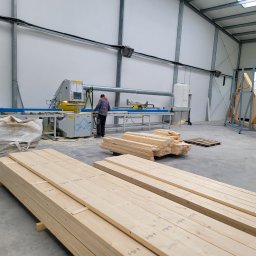 GOLWEST - Produkcja elementów domu szkieletowego: pracownik obsługuje maszynę tnącą drewno w hali. Na pierwszym planie ułożone pakiety desek.