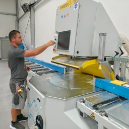 GOLWEST - Pracownik obsługuje precyzyjną piłę panelową Randek w hali. Widoczne elementy sterujące, system odsysania pyłu i obrabiany element z niebieskiego drewna.