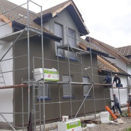 GOLWEST - Budowa domu szkieletowego: elewacja w trakcie prac wykończeniowych, widoczne rusztowania, ocieplenie i ekipa budowlana przy nakładaniu tynku.
