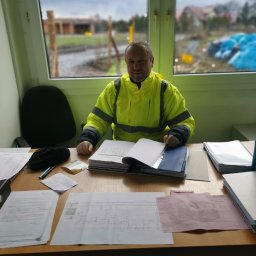 GOLWEST - Mężczyzna w żółtej kurtce przy biurku z dokumentami i planami domu. W tle widok na budowę domu z cegły.