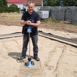 GOLWEST - Pracownik z zagęszczarką wibracyjną na piasku, przygotowując teren pod fundament domu. Widoczne szalunki i ogrodzenie w tle. Prace ziemne.