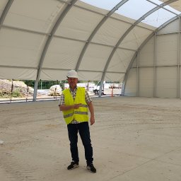 GOLWEST - Wnętrze hali namiotowej z białym dachem łukowym. Mężczyzna w kasku i kamizelce odblaskowej wskazuje na przestrzeń. W tle widać teren budowy.