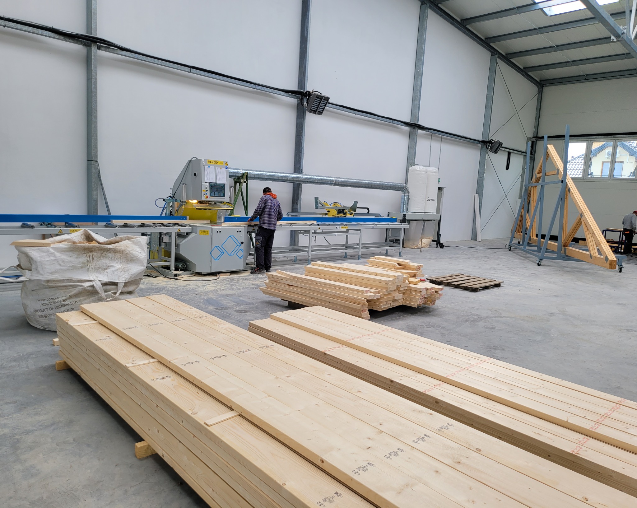 Produkcja elementów domu szkieletowego: pracownik obsługuje maszynę tnącą drewno w hali. Na pierwszym planie ułożone pakiety desek.