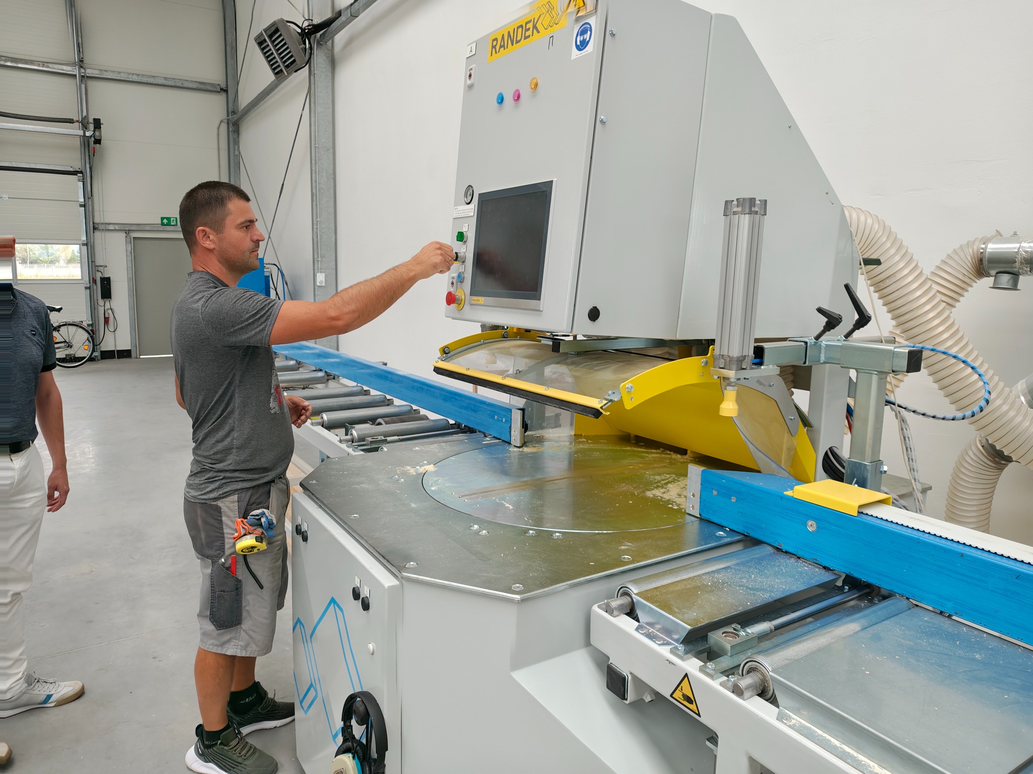 Pracownik obsługuje precyzyjną piłę panelową Randek w hali. Widoczne elementy sterujące, system odsysania pyłu i obrabiany element z niebieskiego drewna.