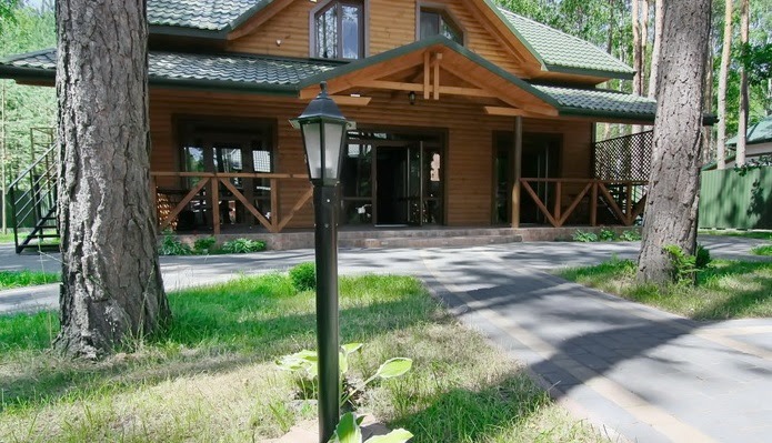 Drewniany dom z zielonym dachem, widoczny z zewnątrz, z lampą ogrodową na pierwszym planie i ścieżką z kostki brukowej prowadzącą do wejścia.