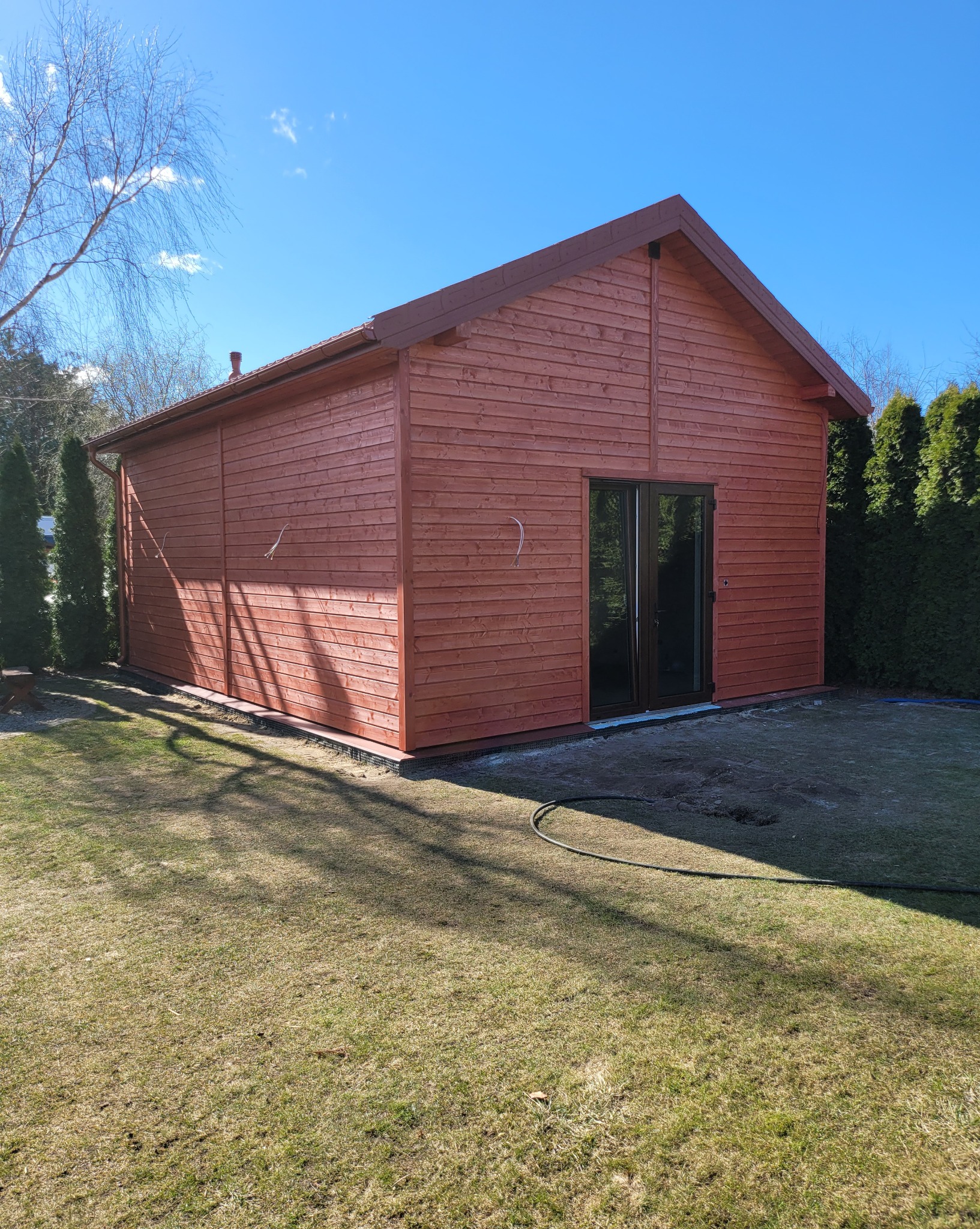 Gotowy, parterowy dom szkieletowy z brązowego drewna, z dwuskrzydłowymi drzwiami tarasowymi, widok z ogrodu w słoneczny dzień, zieleń w tle.