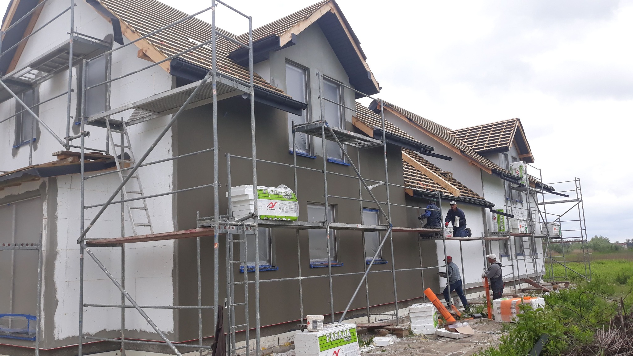 Budowa domu szkieletowego: elewacja w trakcie prac wykończeniowych, widoczne rusztowania, ocieplenie i ekipa budowlana przy nakładaniu tynku.