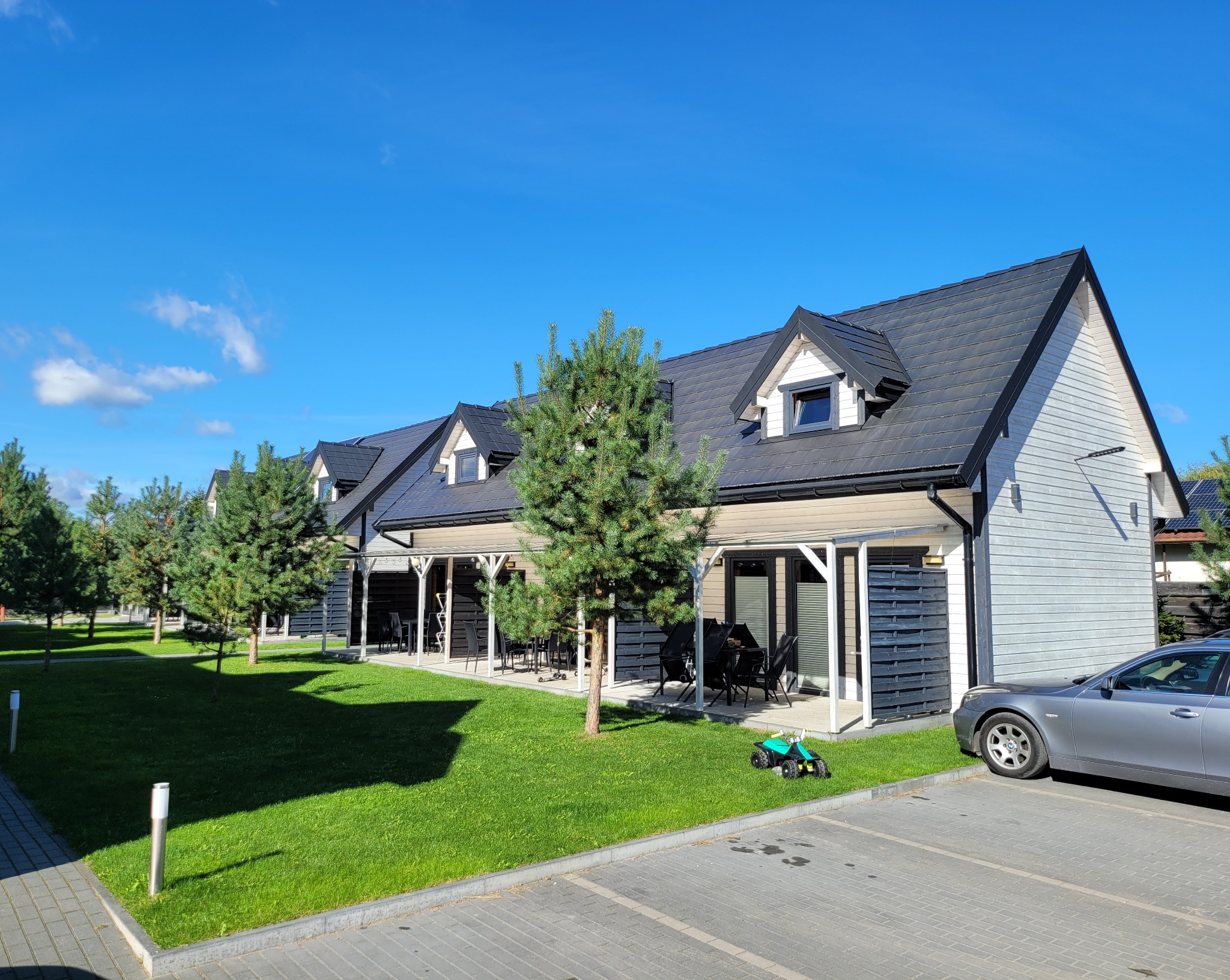 Szereg identycznych domów szkieletowych z czarnymi dachami i jasną elewacją, otoczonych zielenią i zaparkowanym samochodem, w słoneczny dzień.