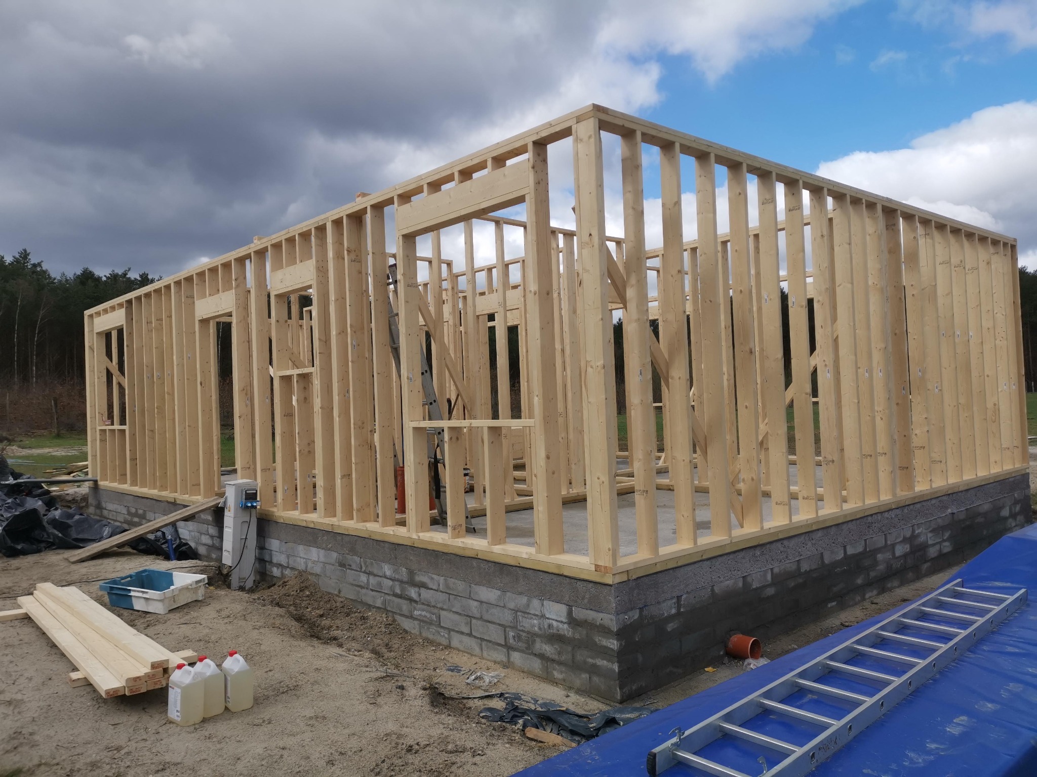 Konstrukcja szkieletowa domu w trakcie budowy. Widoczna drewniana rama, fundament z bloczków, drabina i materiały budowlane. Pochmurne niebo w tle.