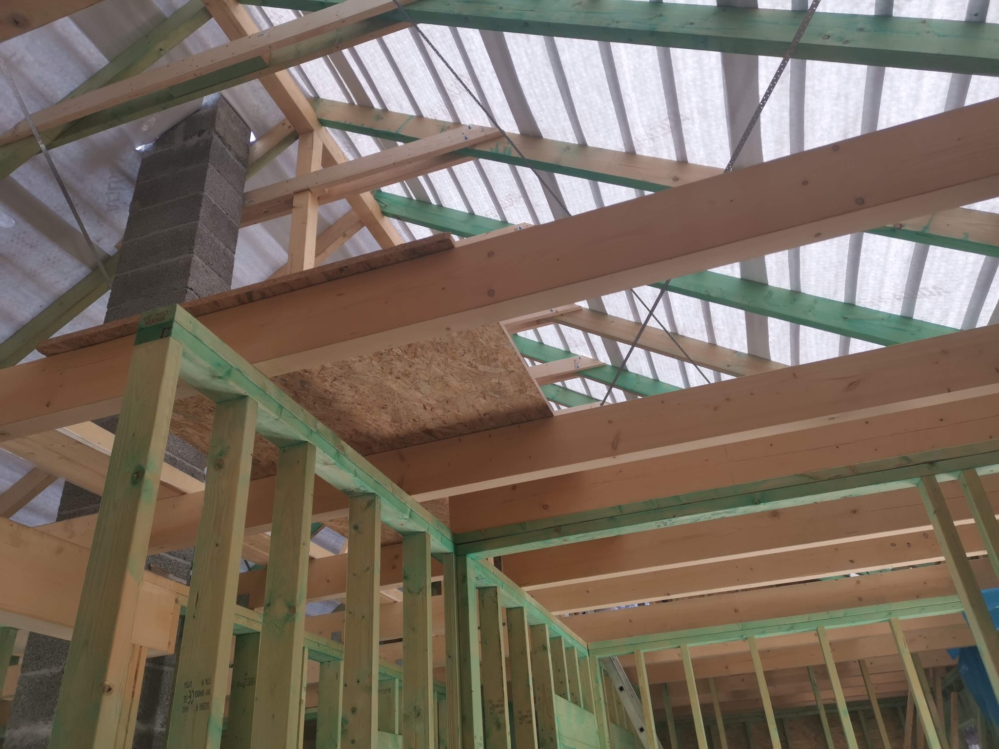 Konstrukcja domu szkieletowego w trakcie budowy: drewniane belki, zielone impregnaty, komin z bloczków betonowych, folia dachowa. Widok od wewnątrz.