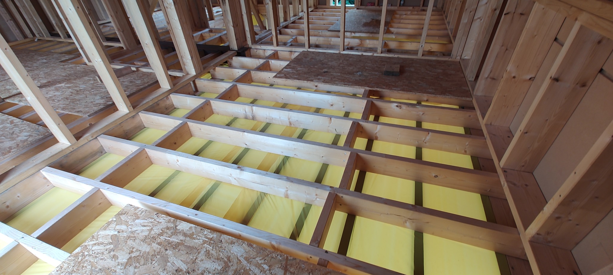 Konstrukcja szkieletowa podłogi z drewnianych belek i żółtą izolacją, widok z góry. Fragment płyty OSB na podłodze. Budowa domu w trakcie realizacji.
