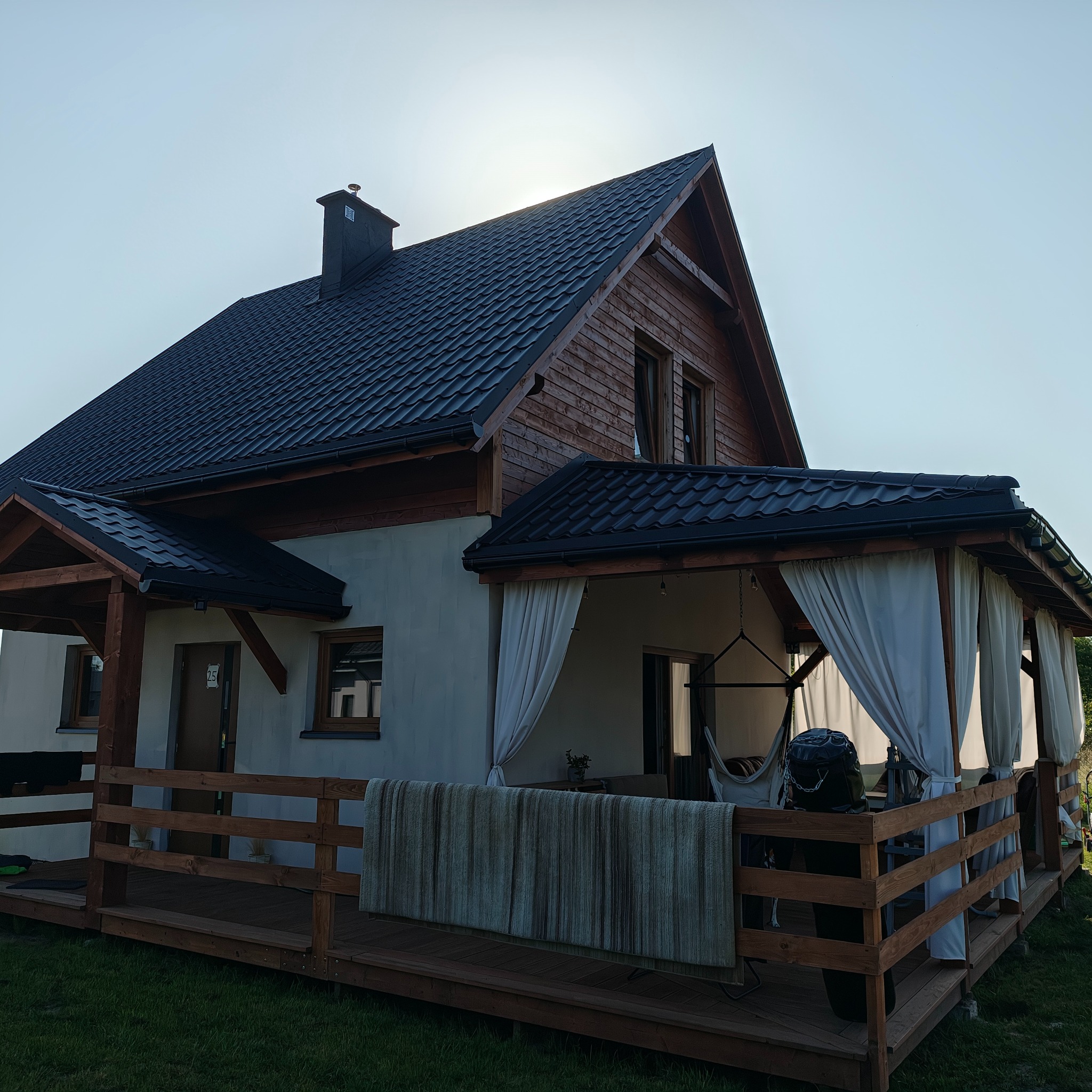 Gotowy dom szkieletowy z ciemnym dachem i drewnianą elewacją. Widoczny taras z zasłonami i hamakiem. Ujęcie z dołu, podkreślające bryłę budynku i jego otoczenie.