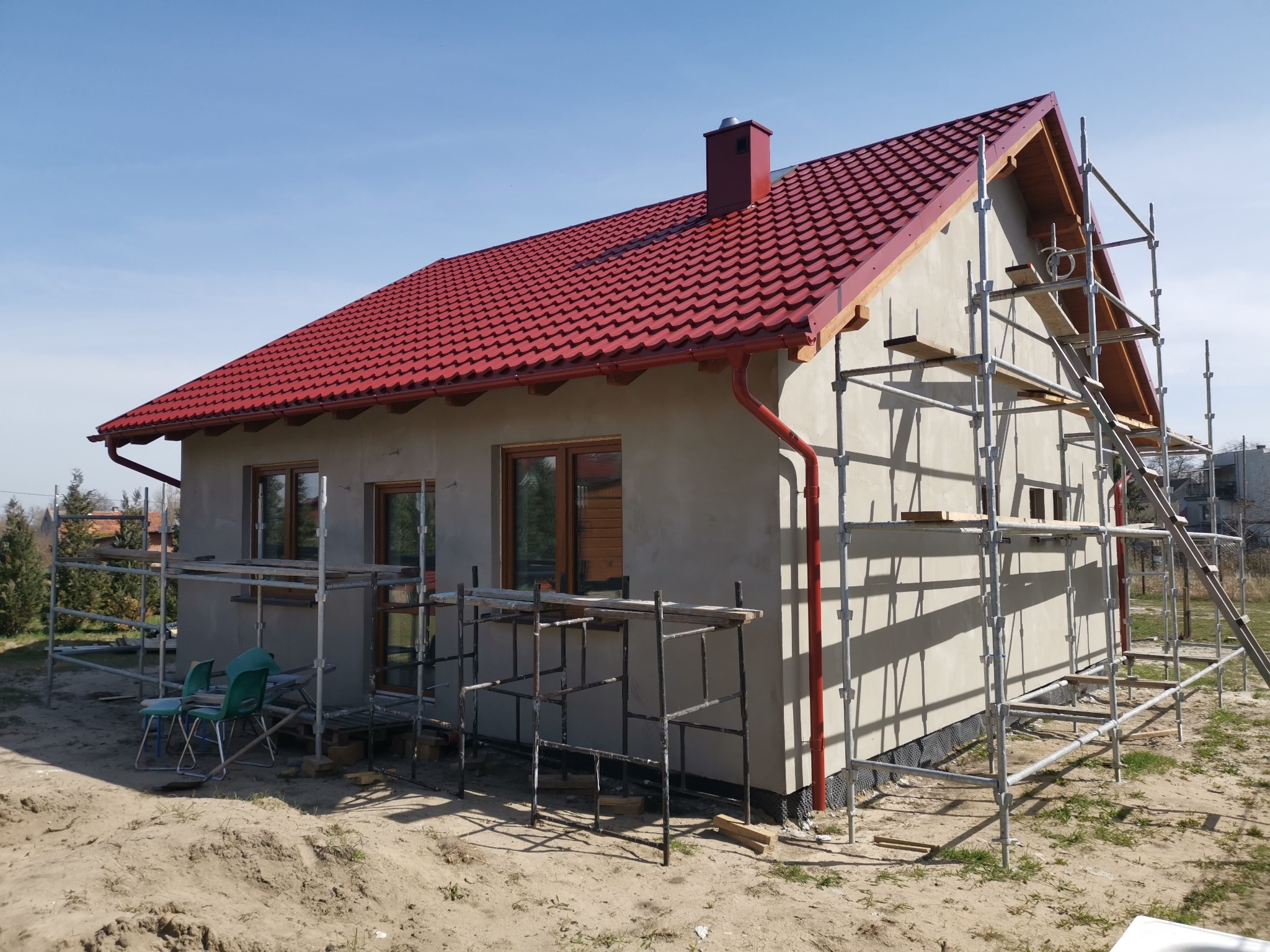 Dom jednorodzinny w trakcie budowy, z czerwonym dachem i rusztowaniami. Widoczne okna i fragment ogrodu. Dzień słoneczny.
