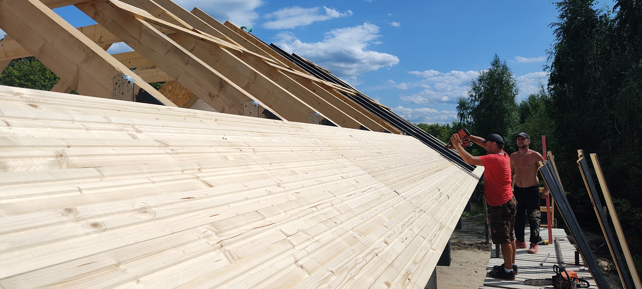 Budowa dachu domu szkieletowego: dwaj mężczyźni montują drewniane deski za pomocą narzędzia pneumatycznego na tle błękitnego nieba i zieleni.