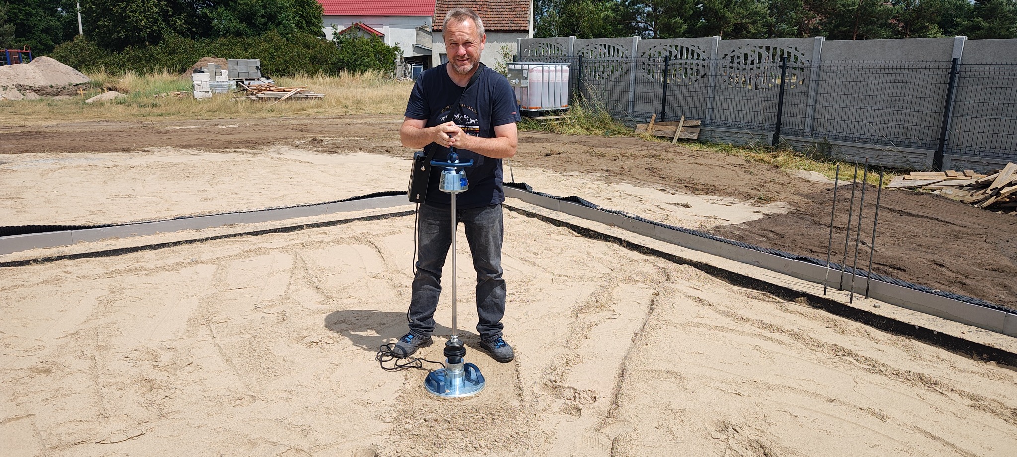 Pracownik z zagęszczarką wibracyjną na piasku, przygotowując teren pod fundament domu. Widoczne szalunki i ogrodzenie w tle. Prace ziemne.
