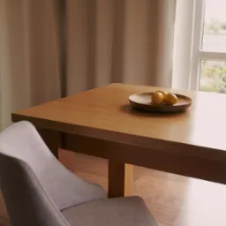 Drewniany stół z owocami na talerzu, obok krzesło z tapicerką w neutralnym odcieniu. W tle okno z zasłoną.