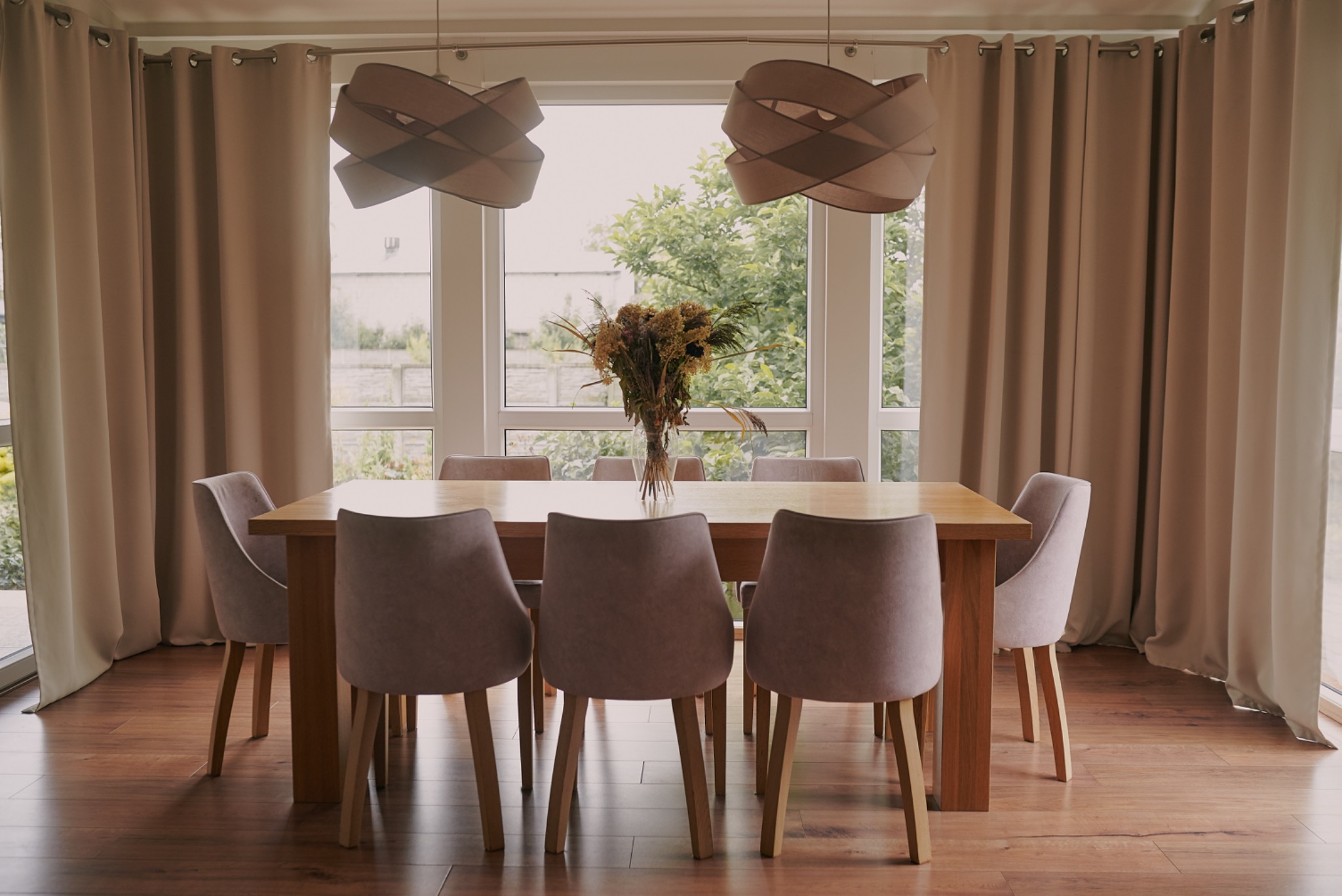 Drewniany stół jadalny z krzesłami w odcieniach beżu, z bukietem w wazonie. W tle okno z widokiem na zieleń i beżowe zasłony. Dwie lampy wiszące nad stołem.
