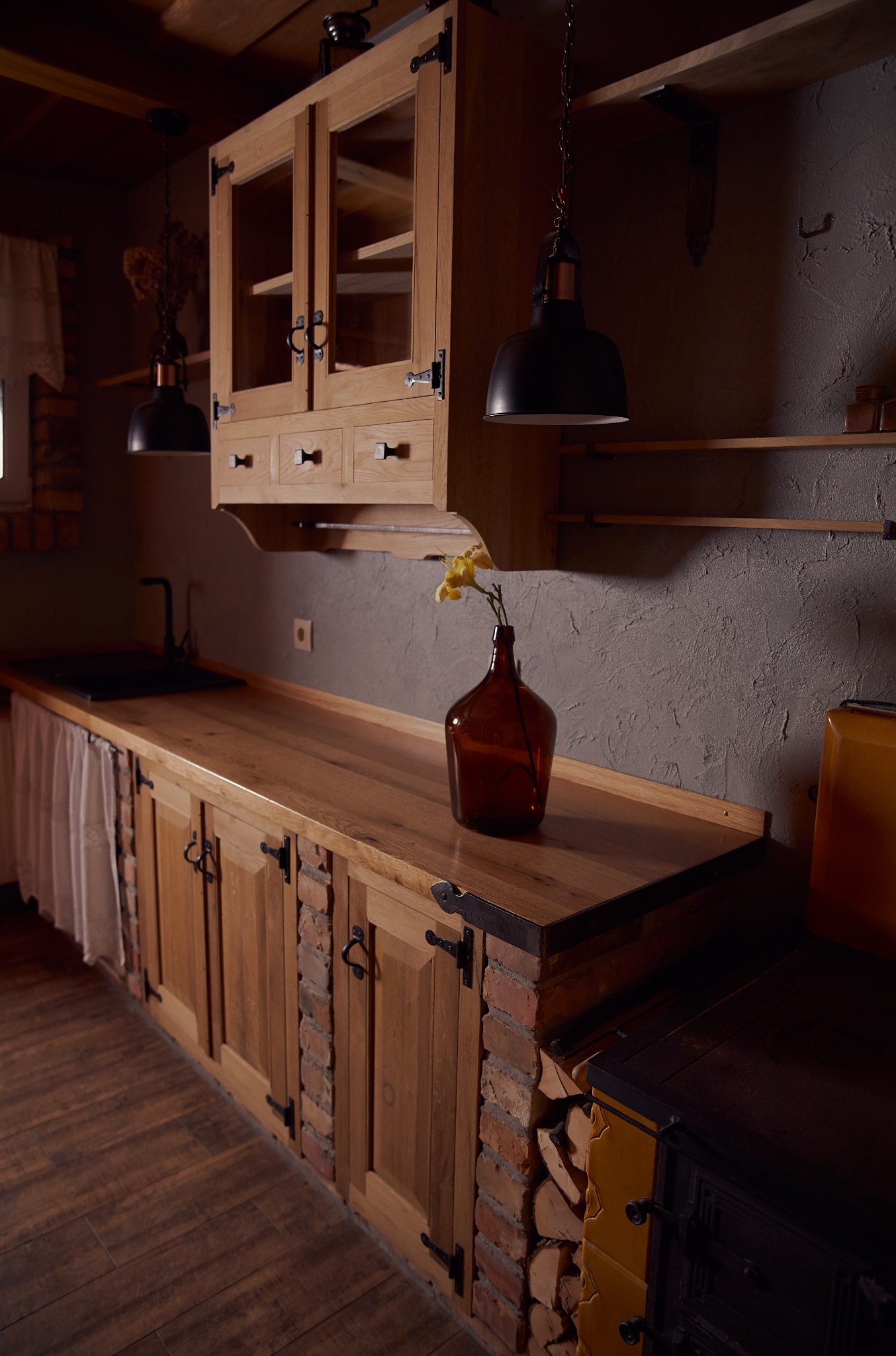 Rustykalna zabudowa kuchenna z drewnianymi szafkami, ceglanymi akcentami i blatem z litego drewna, uzupełniona o lampy w stylu industrialnym i dekoracje.